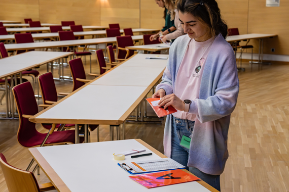Eine Person, die als Testaufsicht arbeitet, steht in einer bestuhlten Halle und bereitet Ihre Unterlagen vor.